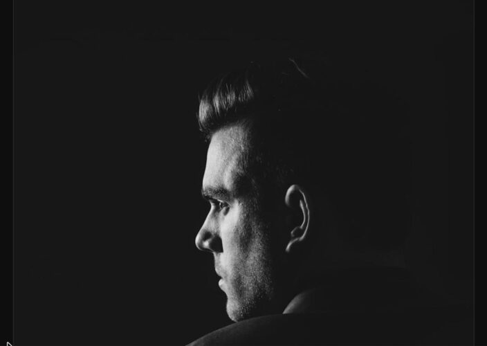 A man in a suit in dramatic black-and-white, his face half-hidden in shadow, evoking the private self-doubt of imposter syndrome